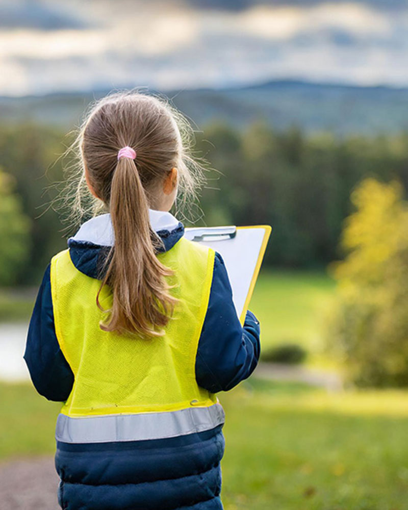  Une jeune fille vêtue d'une veste de haute visibilité tient un presse-papiers et surplombe un champ vert.